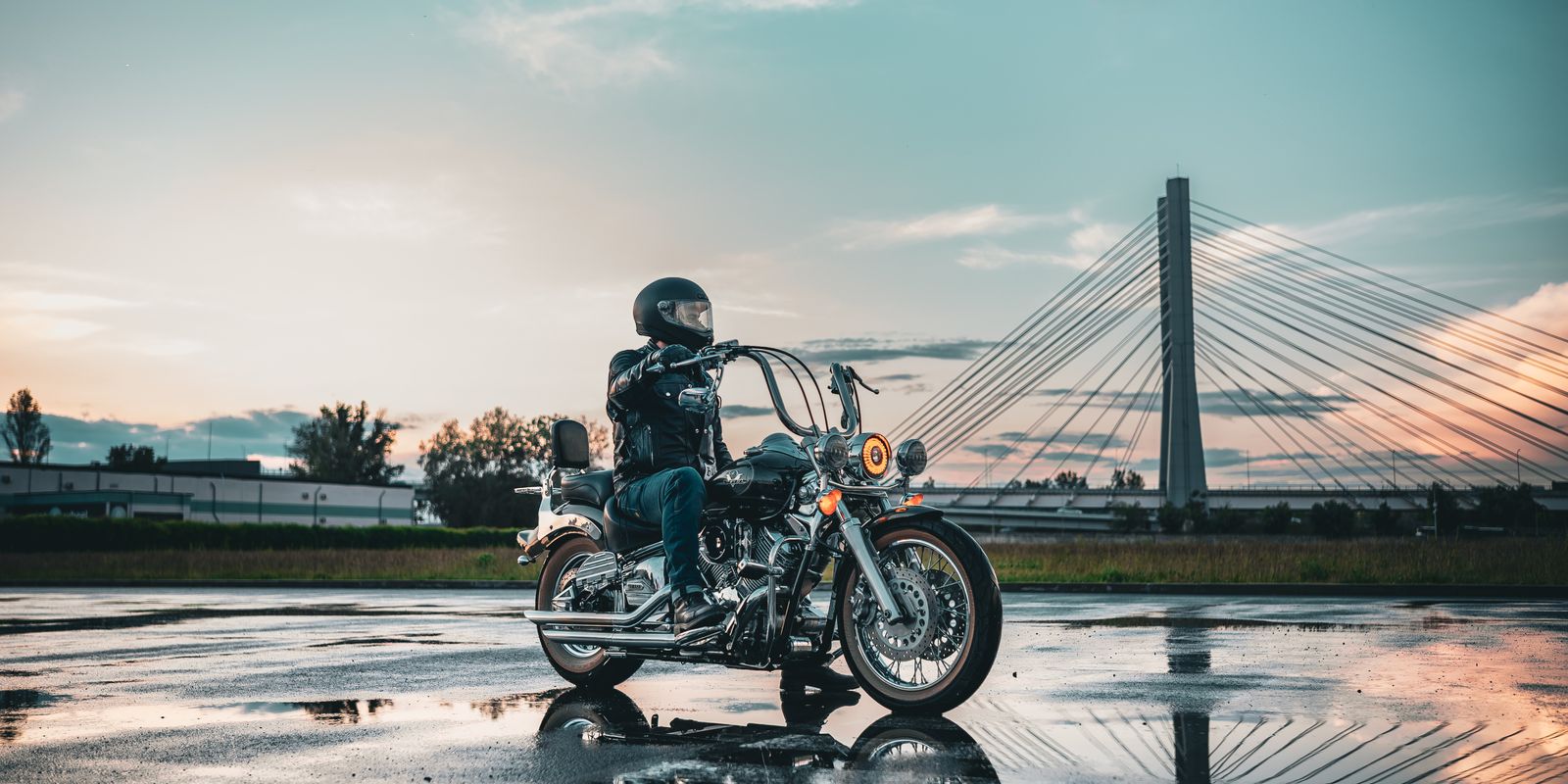 Motoros bőrszerelésben egy vizes úton, háttérben egy híddal naplementekor
