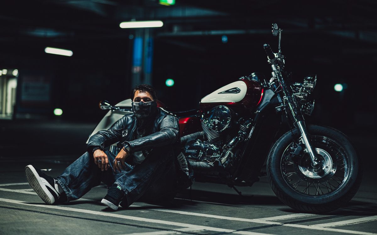 Motoros bőrdzsekiben térdel egy motor mellett egy homályos garázsban