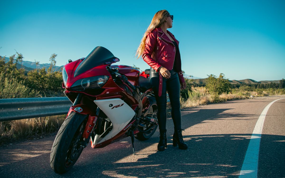 Nő piros bőrdzsekiben egy piros motorkerékpár mellett állva, nyílt úton
